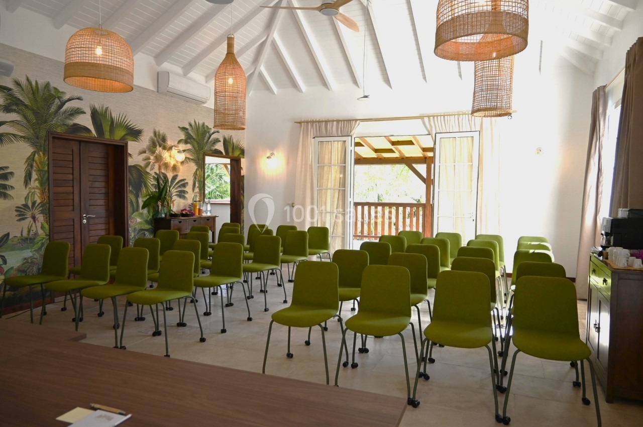 Salle lumineuse aménagée avec des chaises vertes alignées, décor tropical et grandes fenêtres ouvertes sur un balcon.