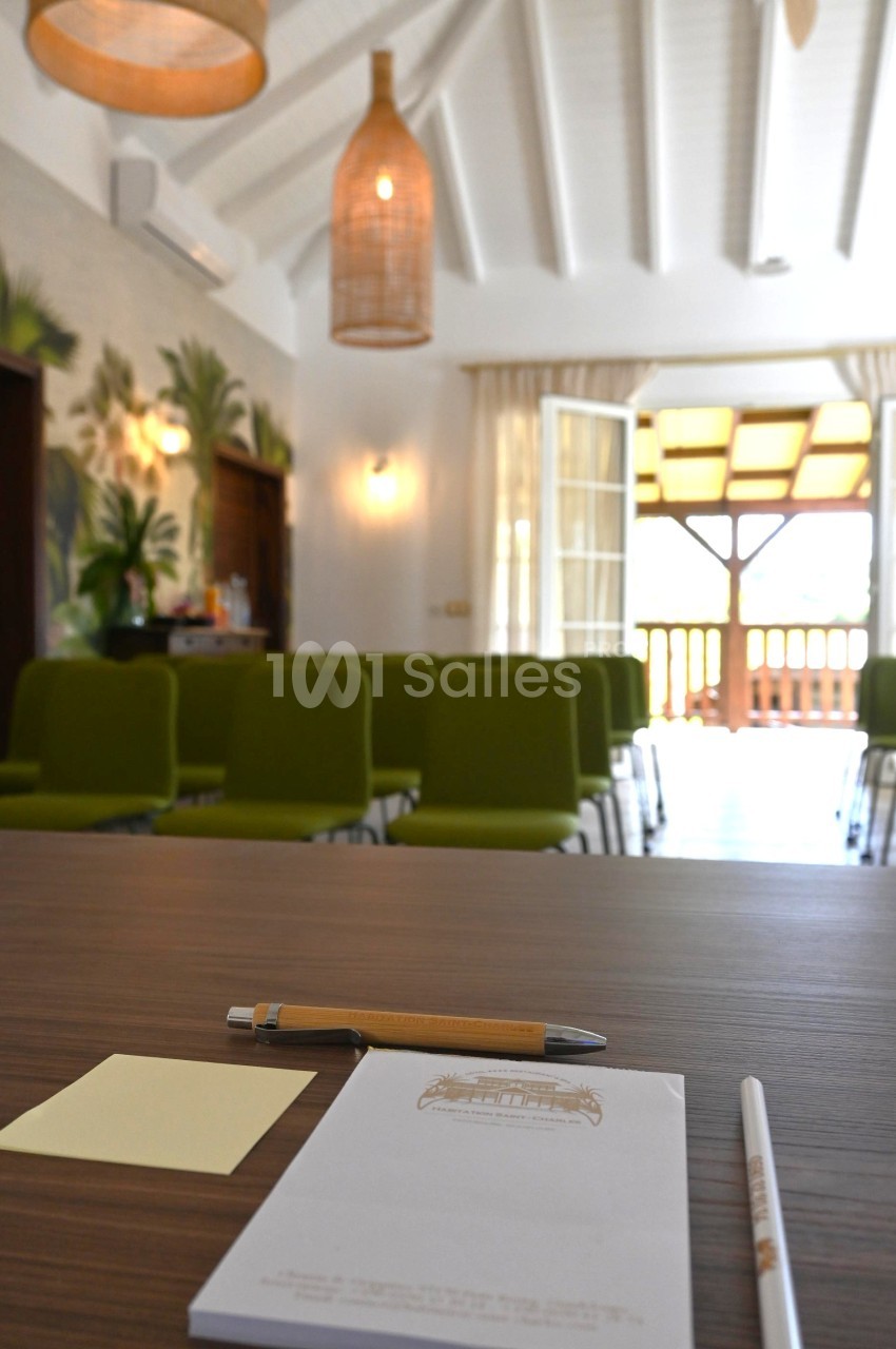 Salle lumineuse avec chaises vertes alignées, vue depuis une table avec bloc-notes, stylo et post-it au premier plan.