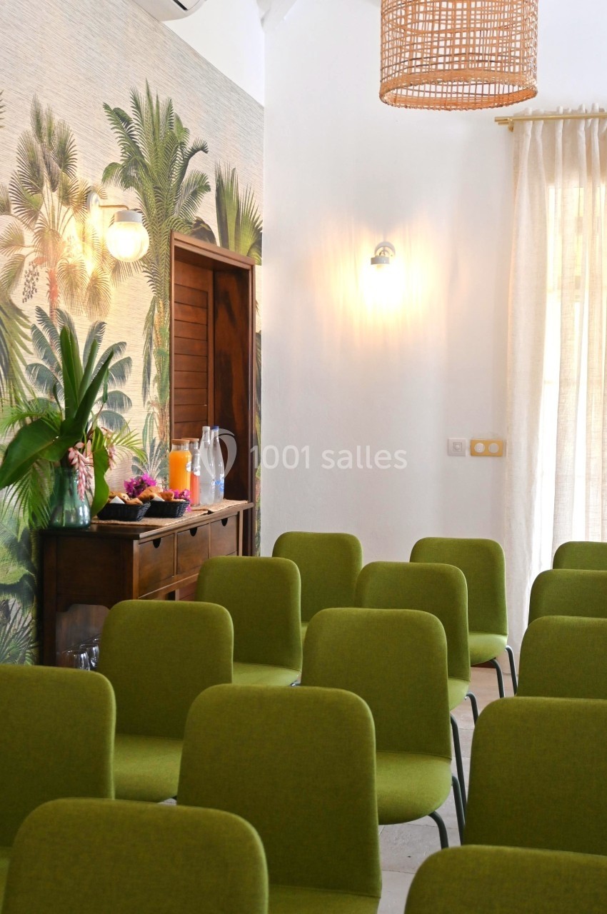 Salle lumineuse avec des chaises vertes alignées, un buffet en bois, des plantes et une suspension en rotin.