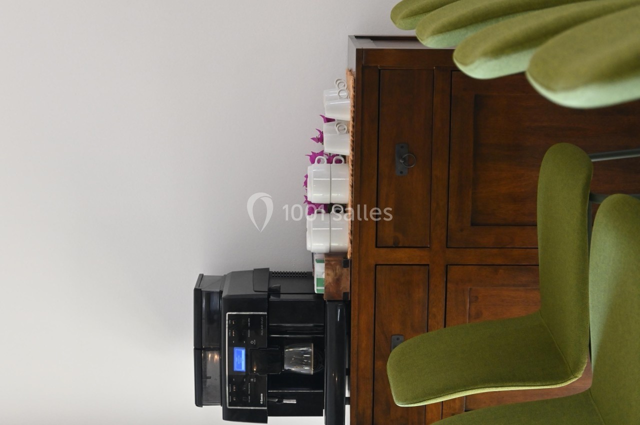 Machine à café noire posée sur un meuble en bois, entourée de tasses blanches et de chaises vertes.