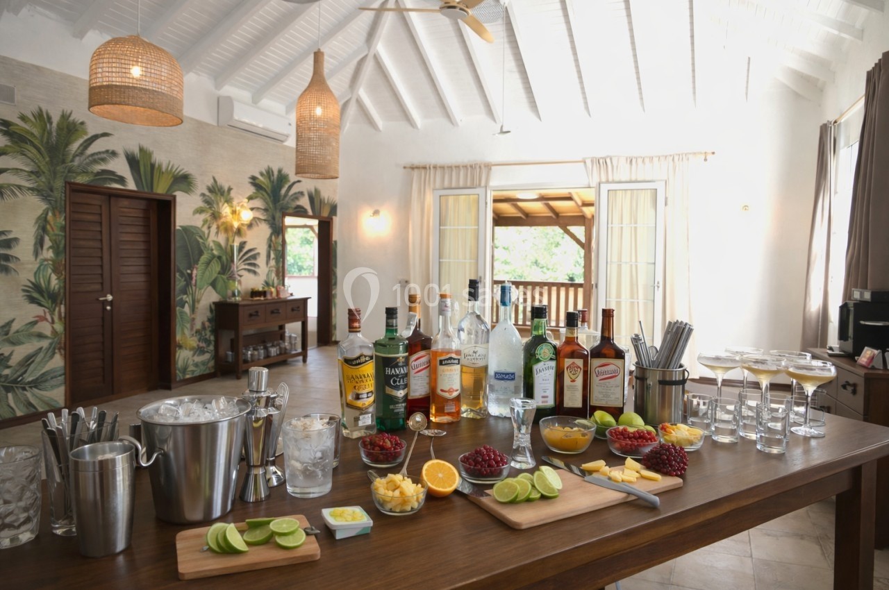Table garnie de bouteilles d'alcool, fruits frais et accessoires de bar dans une pièce lumineuse au décor tropical.