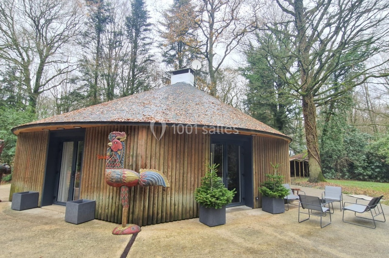 Maison ronde en bois avec toit en métal, entourée d'arbres, décorée d'une sculpture de coq et de plantes en pot.