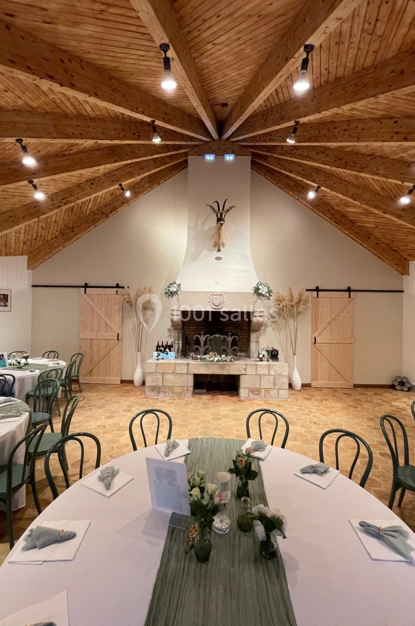 Salle de réception avec tables dressées, cheminée centrale décorée et plafond en bois à poutres apparentes.