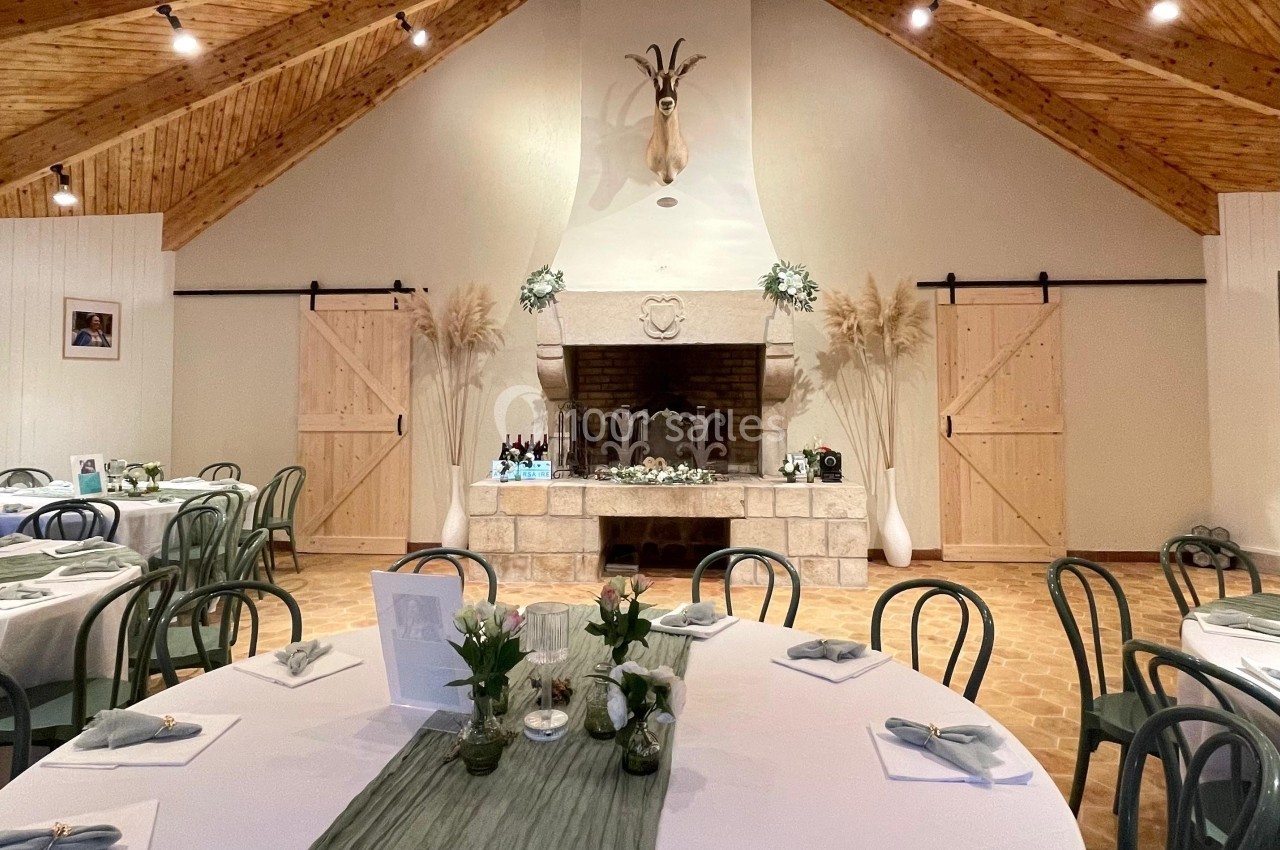 Salle de réception avec tables dressées, cheminée centrale décorée et plafond en bois à poutres apparentes.