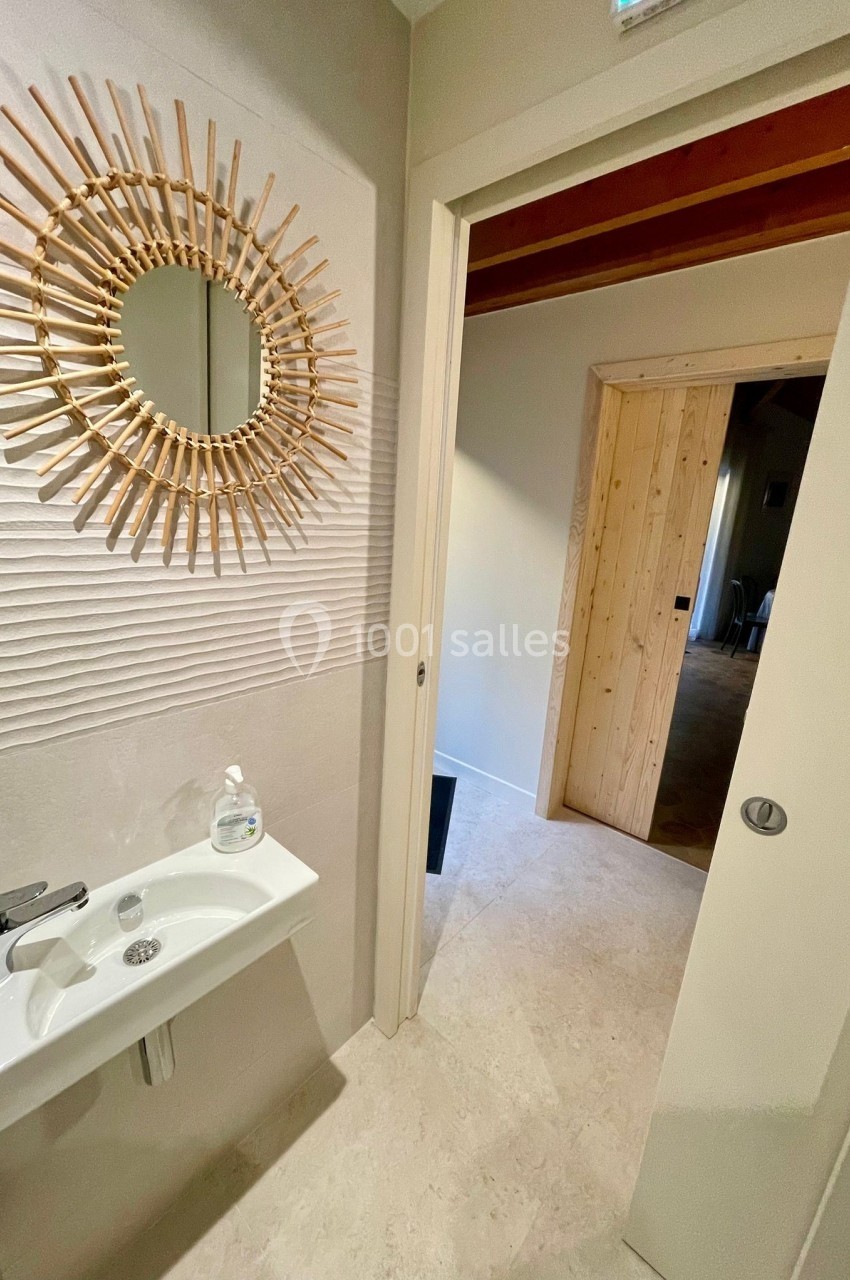 Petit lavabo blanc avec miroir rond en rotin sur un mur texturé, porte ouverte donnant sur un couloir en bois.