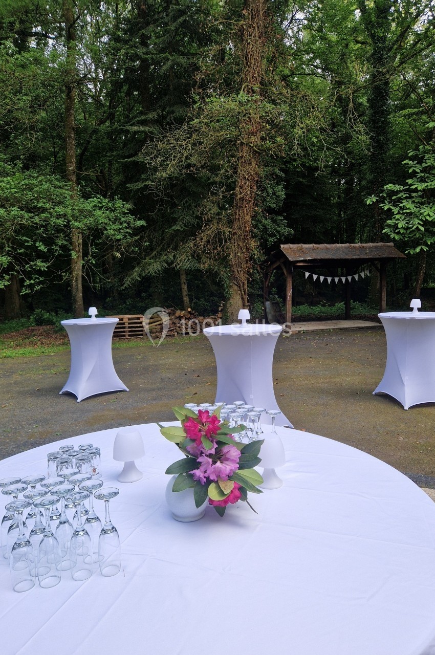 Table ronde dressée avec des verres et un bouquet de fleurs, entourée de mange-debout dans un cadre boisé.