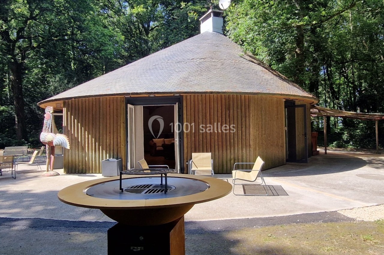 Maison ronde en bois avec toit en chaume, entourée d'arbres, avec mobilier de jardin et barbecue au premier plan.