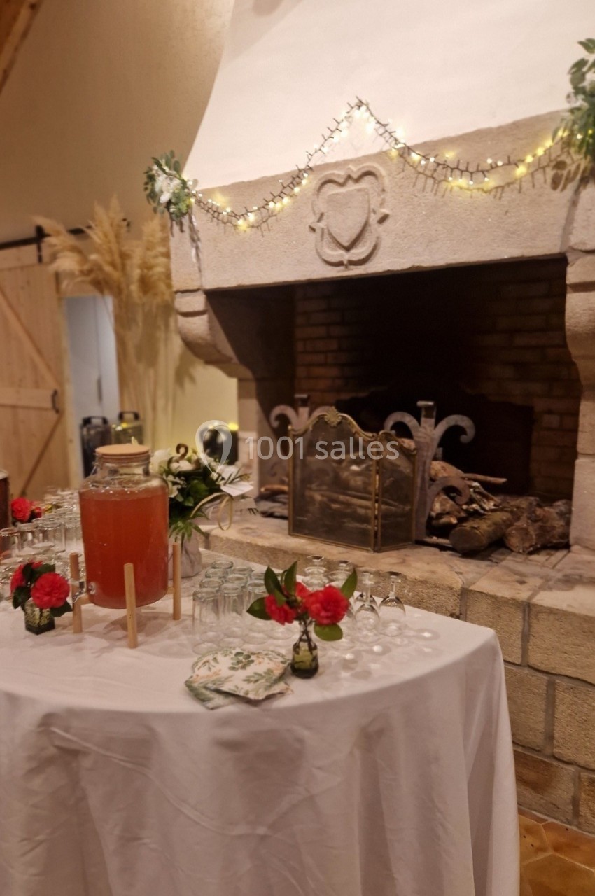 Table décorée avec boissons, verres et fleurs devant une cheminée en pierre ornée de guirlandes lumineuses.