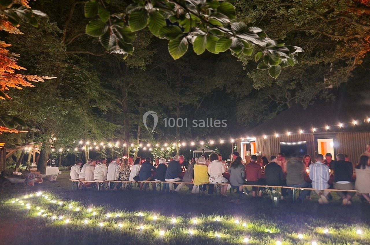 Groupe de personnes assises à une longue table en extérieur, éclairée par des guirlandes lumineuses dans un cadre boisé.