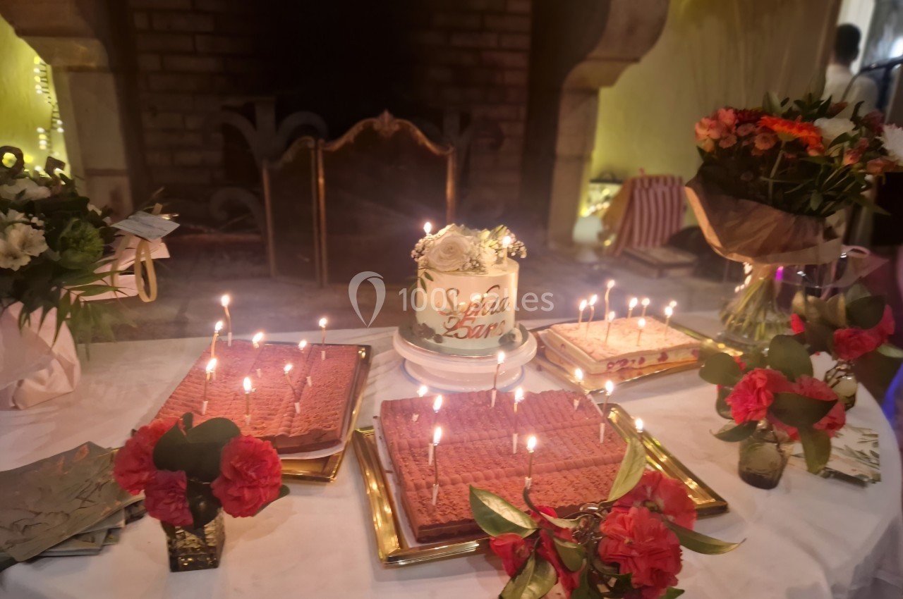 Gâteau décoré et bougies allumées entourés de fleurs et desserts sur une table festive devant une cheminée.