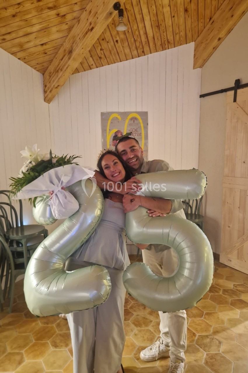 Un couple souriant tenant des ballons argentés formant le chiffre 25 dans une salle à la décoration chaleureuse.