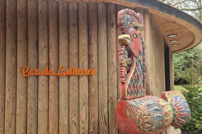 Miniature Enseigne Bois de Guillérien Sculpture colorée en forme d'oiseau devant un bâtiment en bois avec l'inscription ’Bois des Guillemins’.