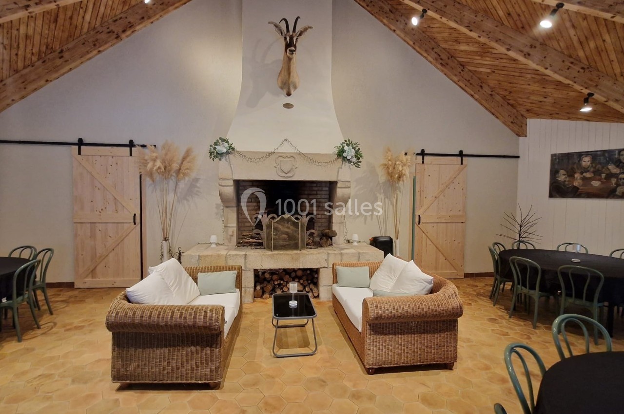 Salle avec cheminée centrale, fauteuils en rotin, tables rondes et plafond en bois sous une tête de cerf décorative.