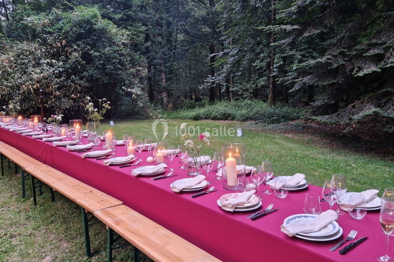 Grande table dressée avec une nappe rouge, couverts et bougies, installée en extérieur dans un cadre boisé.