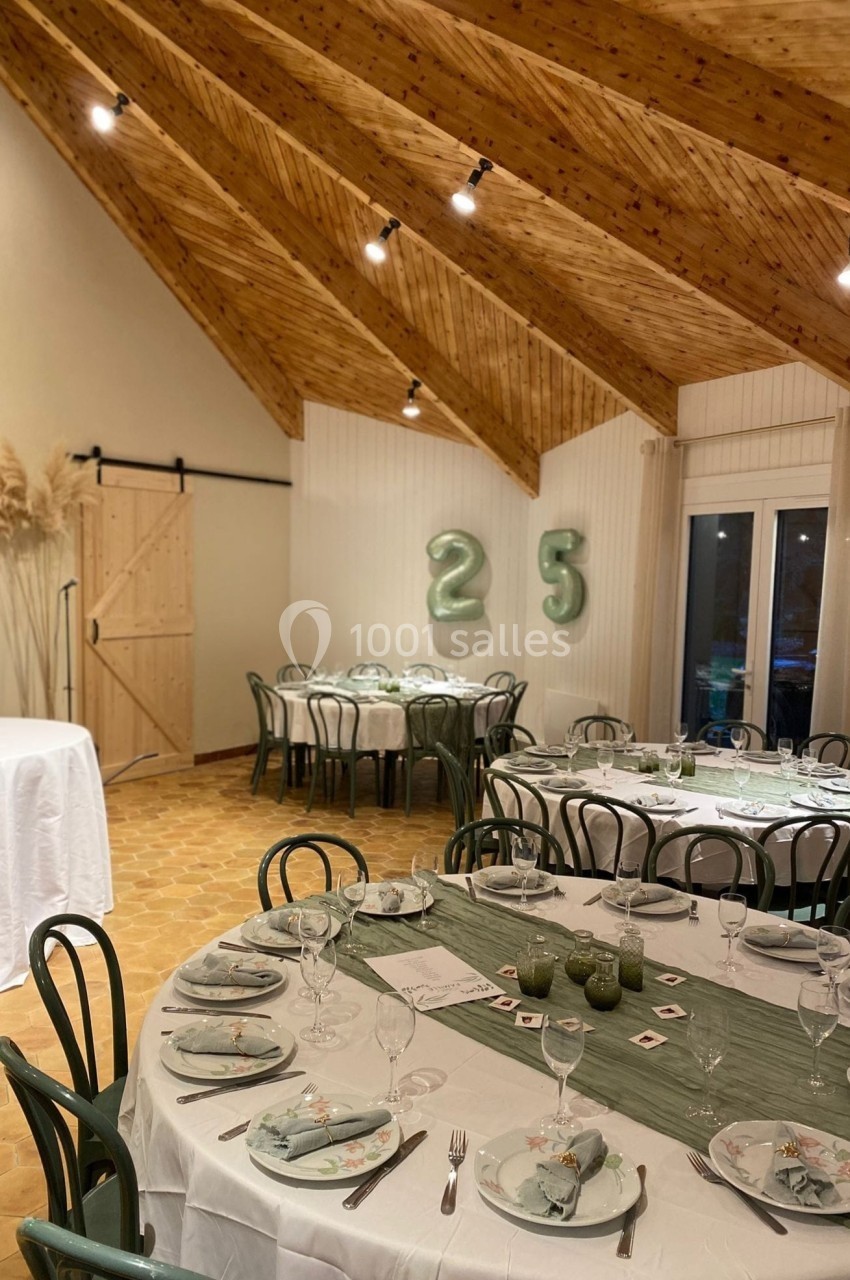 Salle décorée pour une fête avec tables dressées, chaises vertes, ballons ’25’ et plafond en bois.