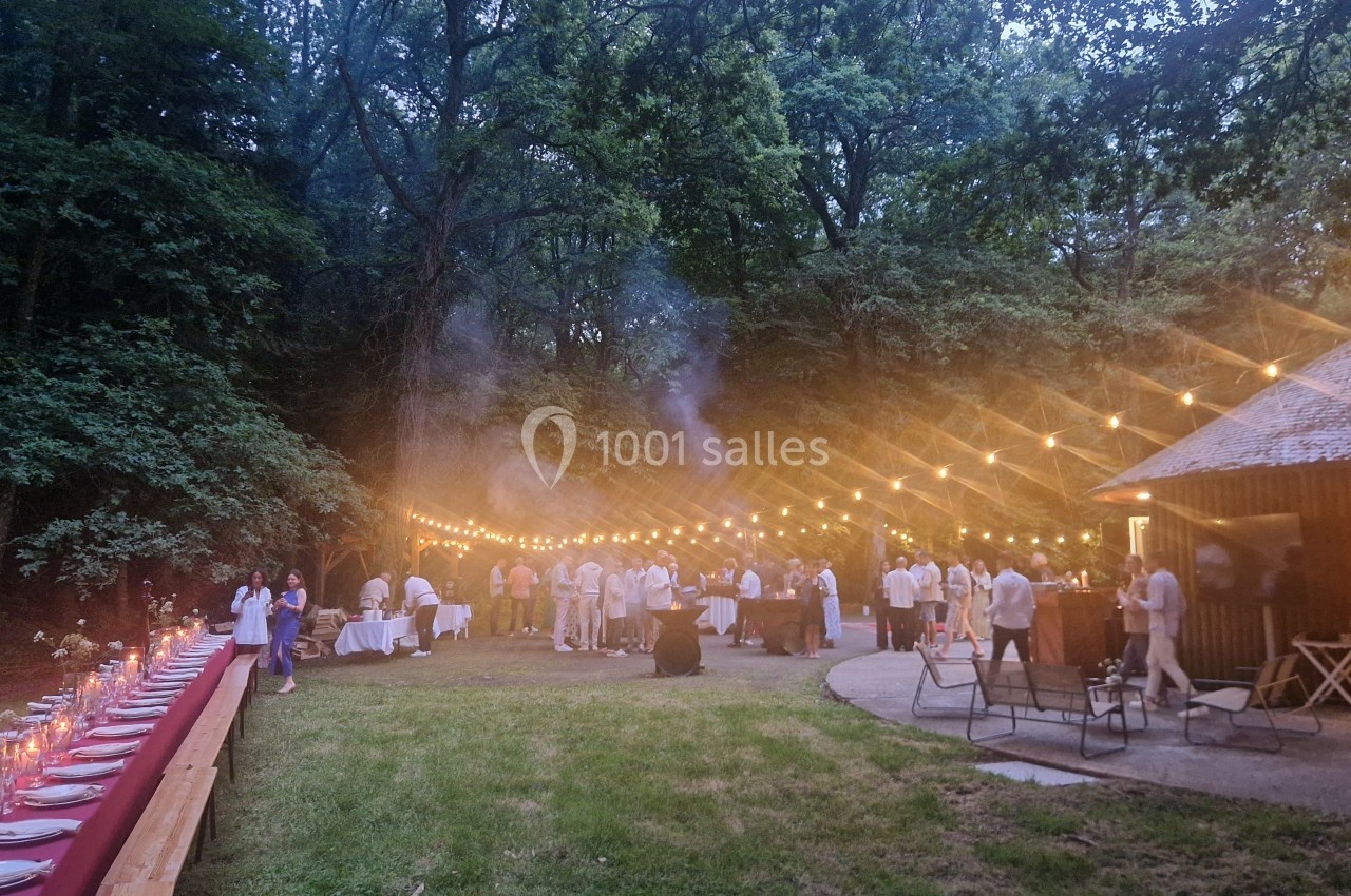 Dîner en plein air sous des guirlandes lumineuses, avec des invités rassemblés près d'un bâtiment en bois dans un cadre…