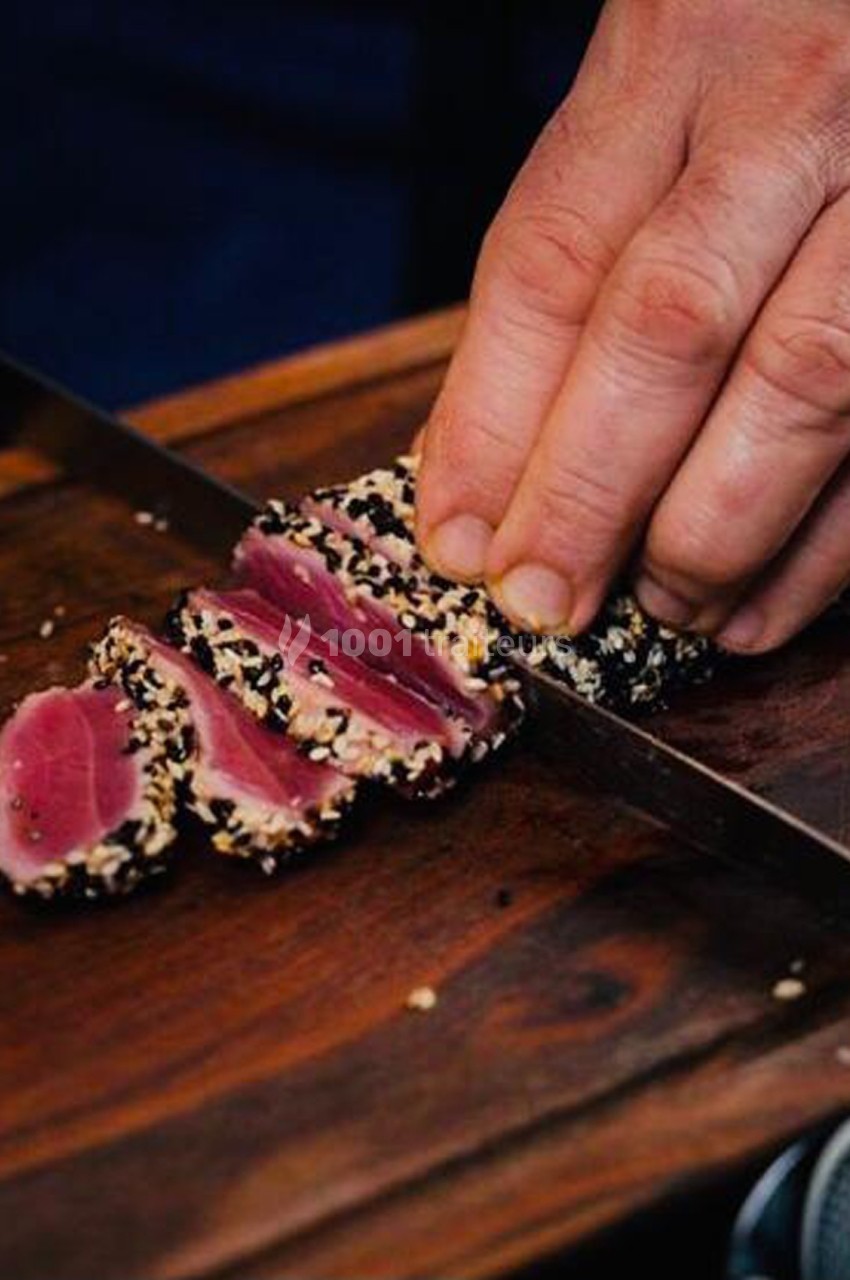 Main coupant un filet de thon mi-cuit en tranches, enrobé de graines de sésame, sur une planche en bois.