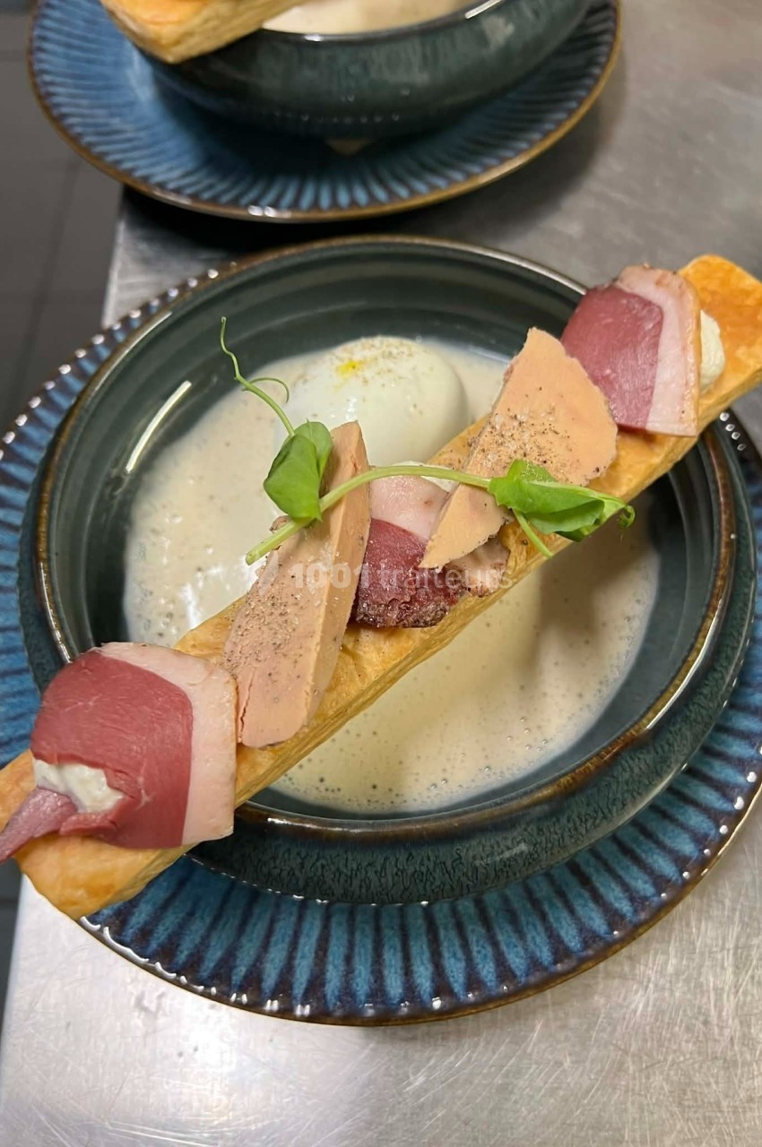 Assiette contenant une soupe crémeuse, surmontée d'une pâte feuilletée garnie de foie gras, magret fumé et pousses vertes.