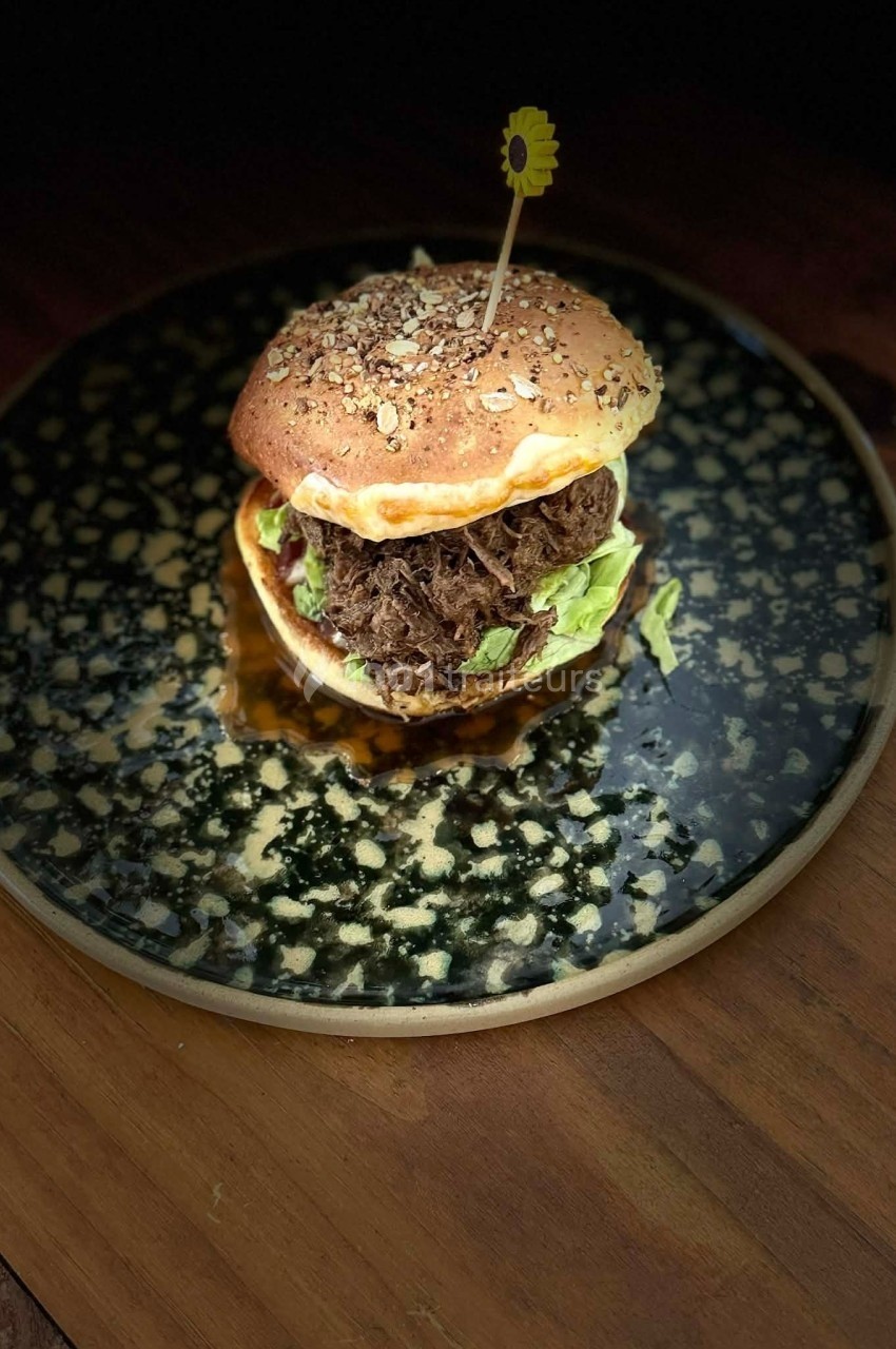 Burger garni de viande effilochée, salade et sauce, présenté sur une assiette noire avec un cure-dent décoratif.