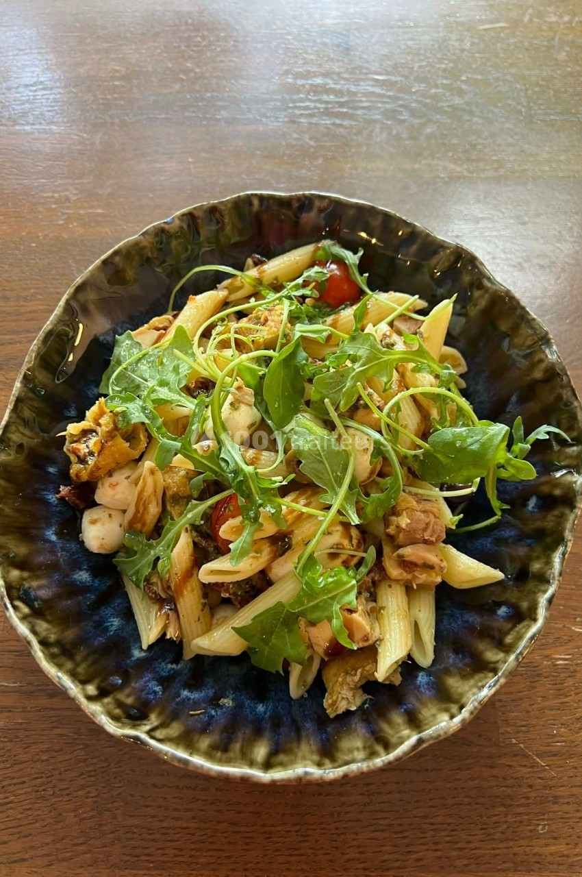 Assiette de pâtes garnie de roquette, tomates cerises, morceaux de poulet et fromage, servie dans un bol en céramique.