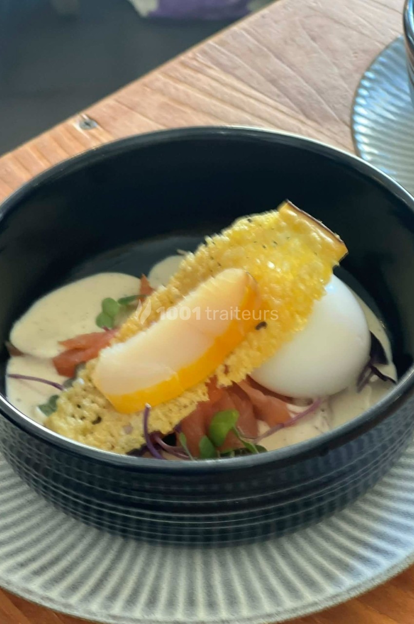 Œuf poché avec saumon fumé, crème et croustillant de fromage, servi dans un bol noir sur une assiette texturée.