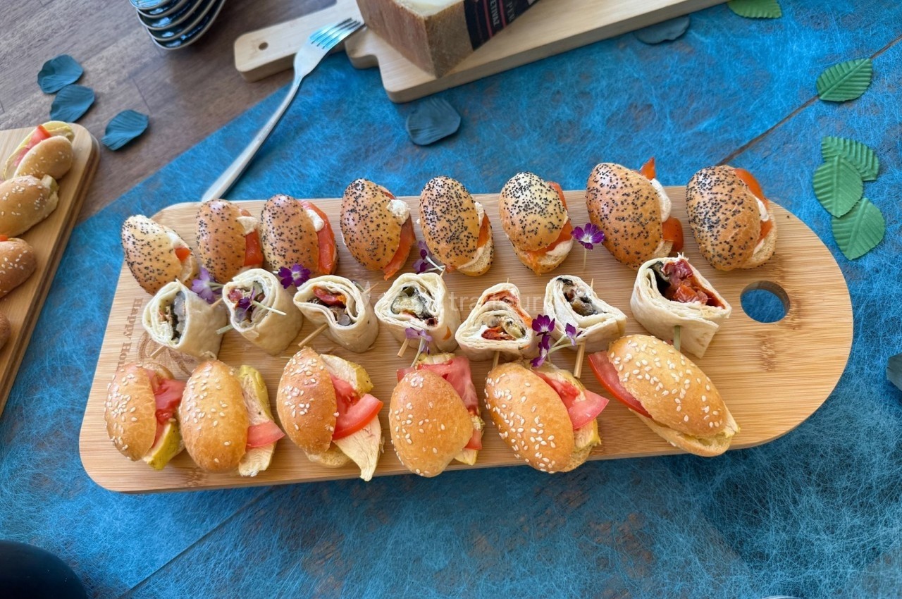 Plateau en bois avec mini-sandwichs garnis et wraps roulés, disposés sur une nappe bleue avec des feuilles décoratives.
