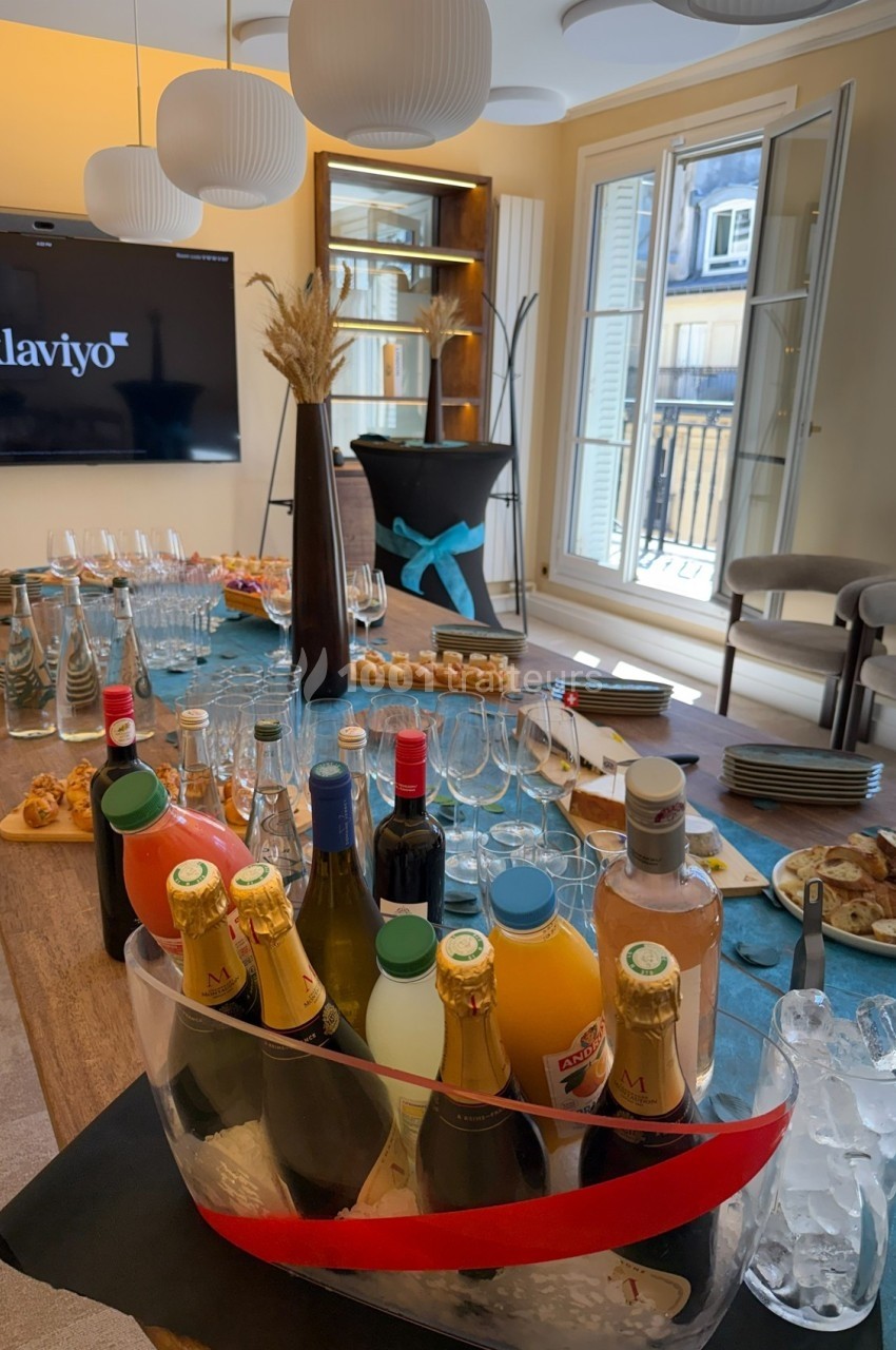 Table dressée avec boissons, verres, viennoiseries et snacks dans une salle lumineuse avec écran et grandes fenêtres.