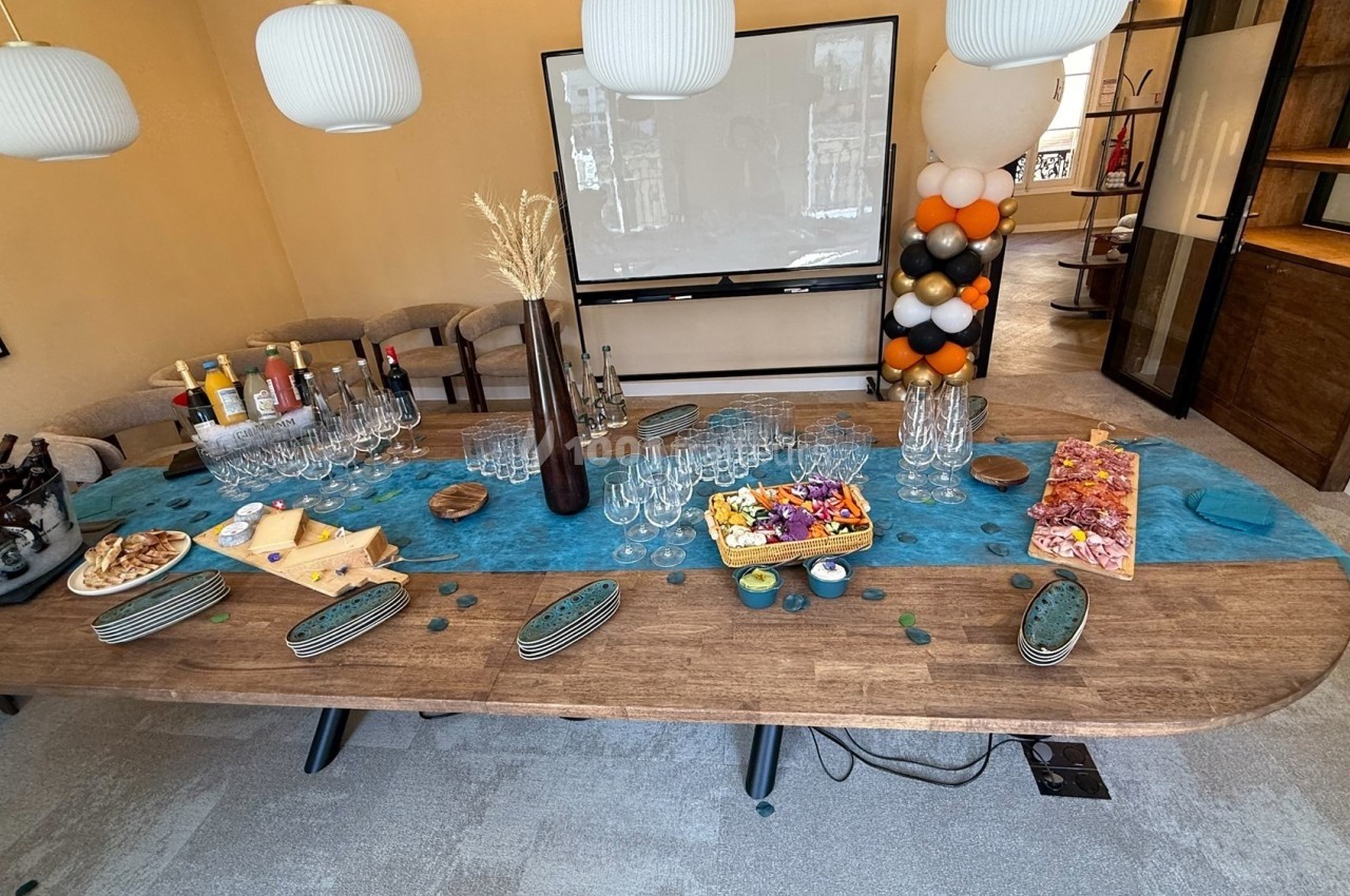 Table dressée avec des boissons, verres, plateaux de charcuterie et apéritifs, dans une salle lumineuse avec écran et…