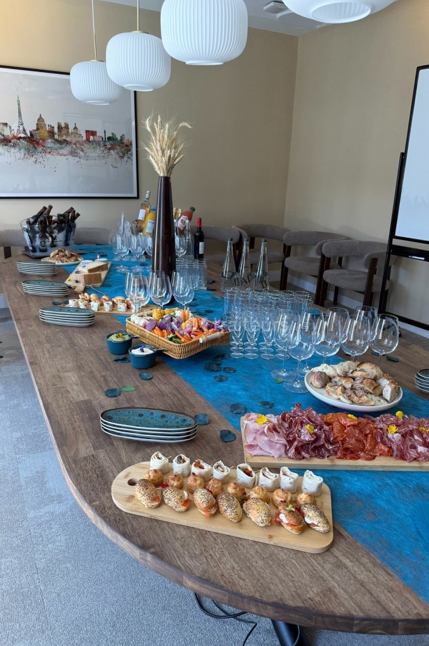 Table garnie de mets variés, verres à vin et décorations, dans une salle lumineuse avec un tableau en arrière-plan.