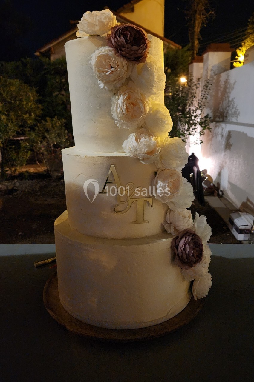 Gâteau de mariage à trois étages décoré de fleurs blanches et violettes, avec des initiales dorées ’A & T’.