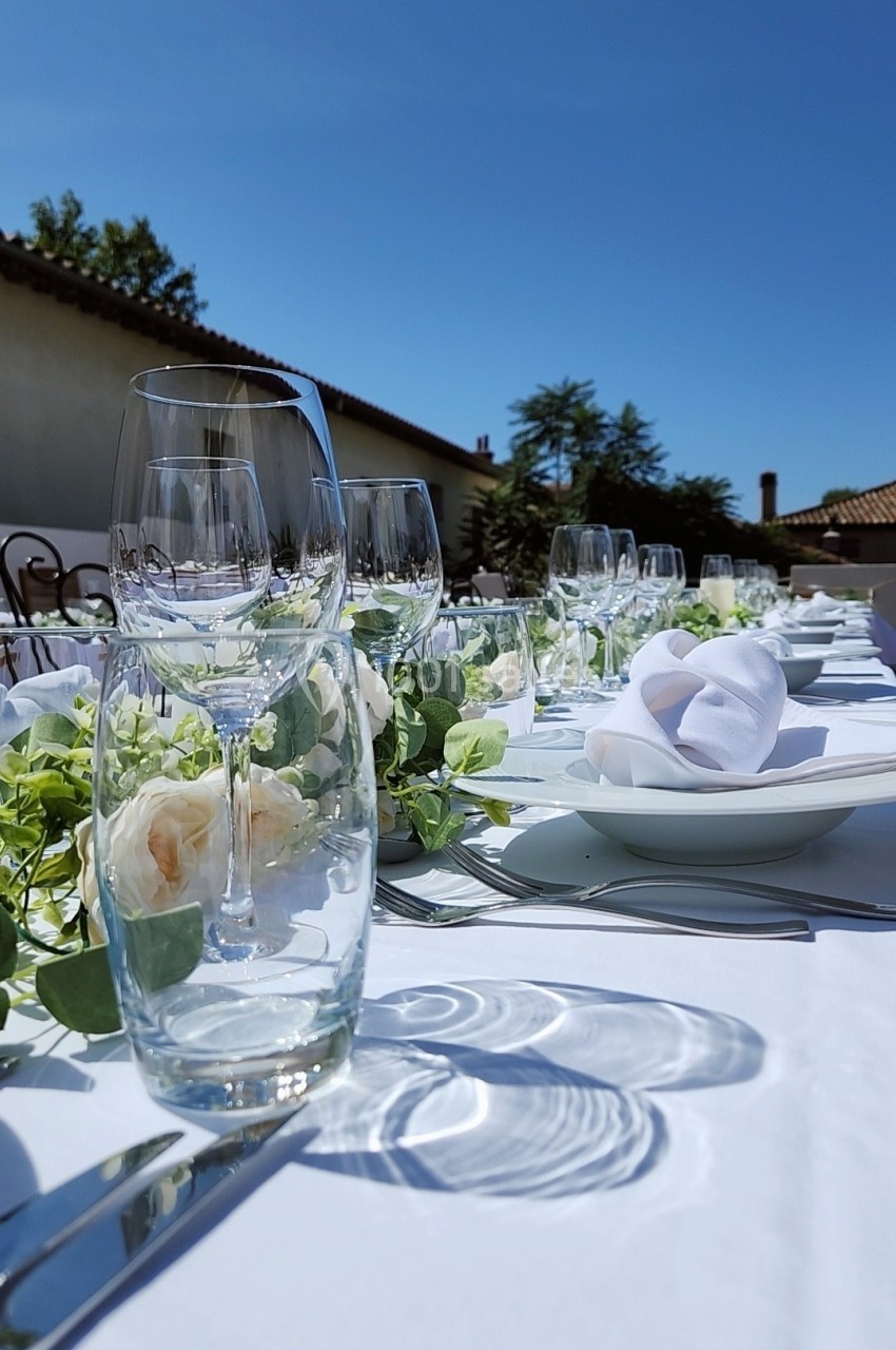 Table dressée en extérieur avec verres, assiettes, serviettes pliées et décoration florale sous un ciel bleu.