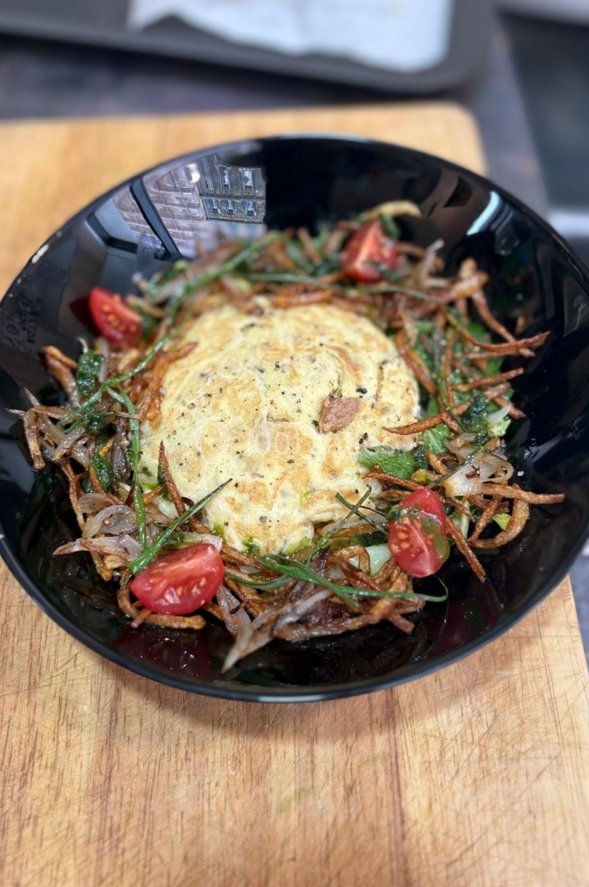 Omelette garnie de tomates cerises, oignons frits et herbes fraîches, servie dans un bol noir sur une planche en bois.