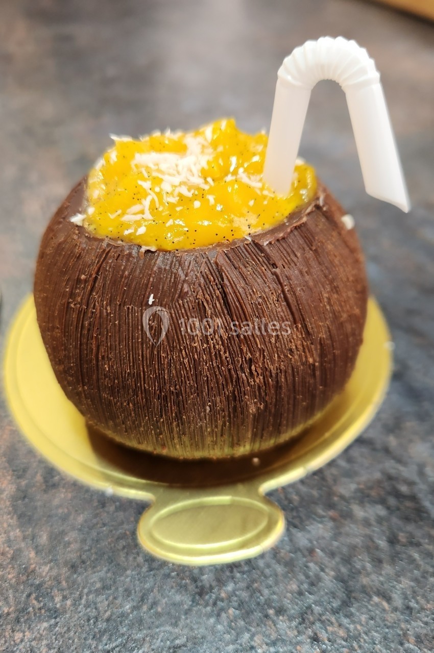Dessert en forme de noix de coco, garni de mousse jaune et de copeaux, avec une paille blanche décorative.