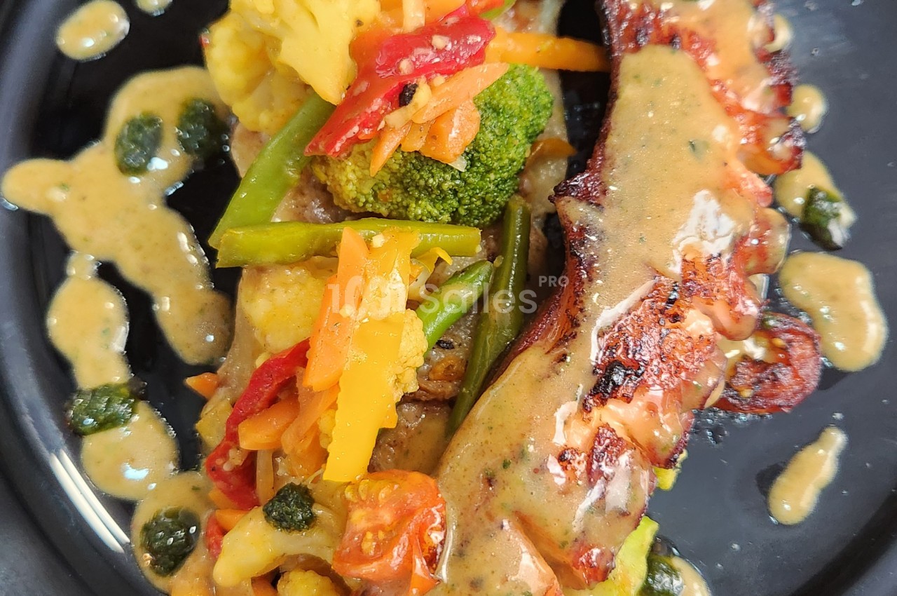 Assiette noire garnie de légumes colorés (chou-fleur, brocoli, poivron, carotte, haricots verts) et de tomates cerises, accompagnée d'une viande rôtie nappée d'une sauce crémeuse.