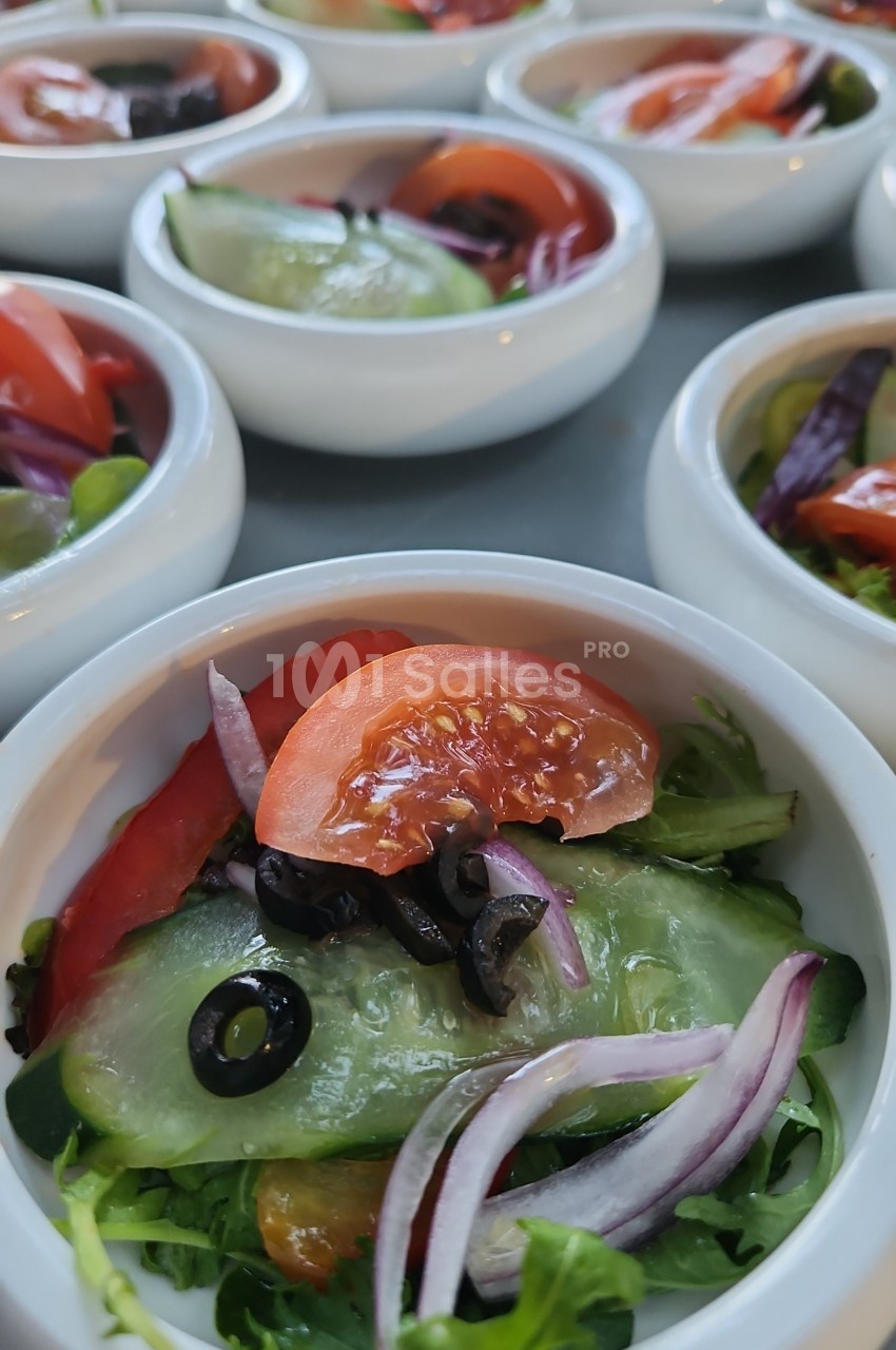 Petites coupelles blanches de salade composée : tomates, concombre, oignon rouge, olives noires et feuilles vertes, dressées en rangées.