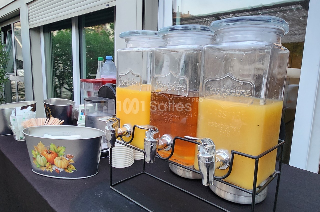Trois distributeurs en verre de jus d'orange et de boisson ambrée sur une table sombre, gobelets empilés à côté.