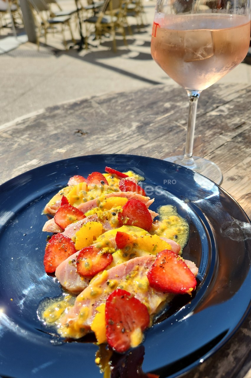 Assiette bleue de tranches de poisson cru nappées de sauce jaune à la passion, garnie de fraises et de quartiers d'orange, avec un verre de rosé en arrière-plan.