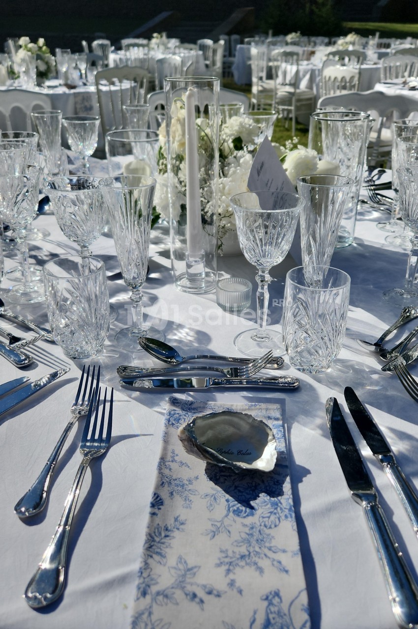 Table dressée en extérieur : nappe blanche, verres en cristal, couverts argentés, serviette bleue et huître posée dessus.