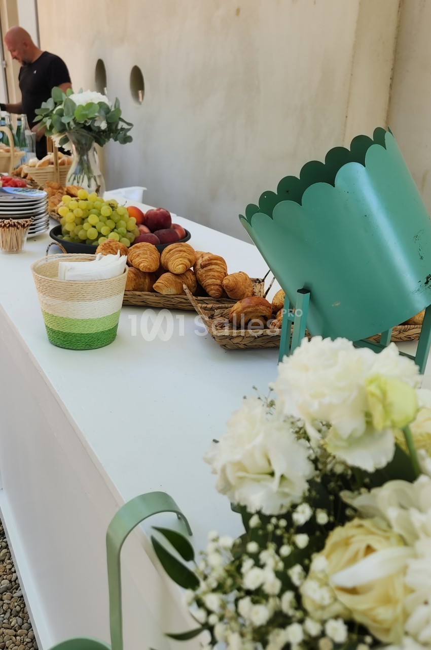 Buffet extérieur sur table blanche avec croissants, raisins verts, pommes, corbeilles en osier, assiettes empilées et bouquets de fleurs blanches.