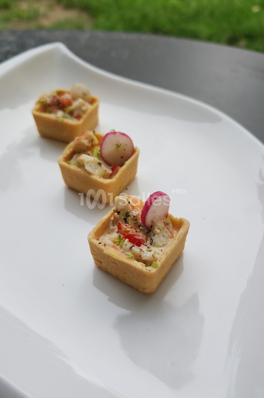 Trois petites tartelettes carrées à pâte dorée garnies d'une préparation de fruits de mer et de légumes, surmontées d'une tranche de radis, posées sur une assiette blanche.