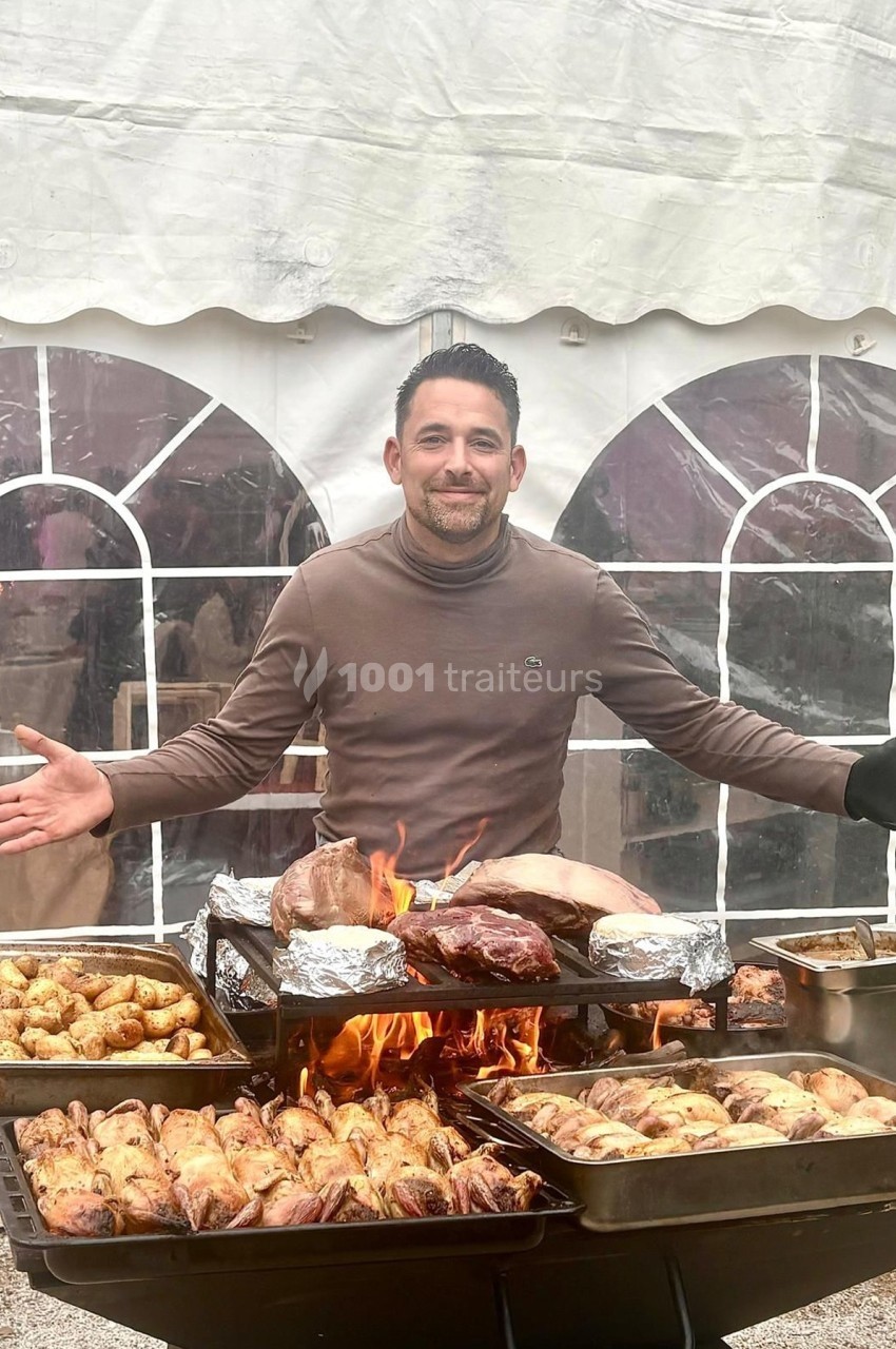 Un homme souriant devant un barbecue chargé de viandes et de pommes de terre, sous une tente blanche.