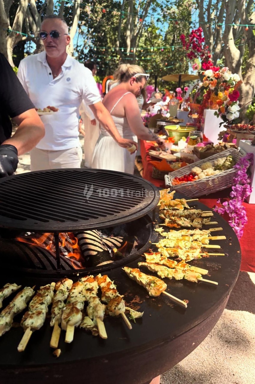 Grill circulaire avec brochettes de poulet en cuisson, buffet coloré en arrière-plan dans un cadre extérieur ensoleillé.