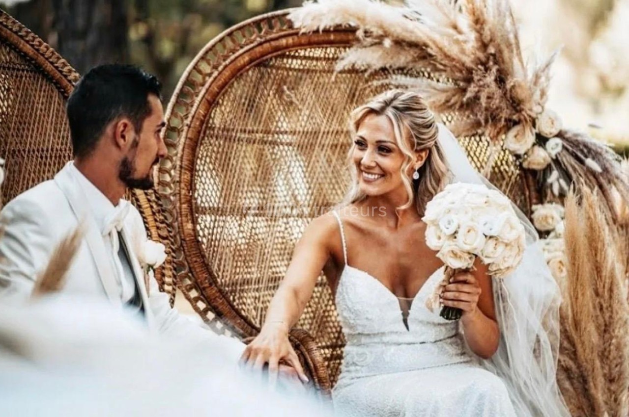 Une mariée souriante en robe blanche tient un bouquet de fleurs, assise à côté d'un homme en costume clair.