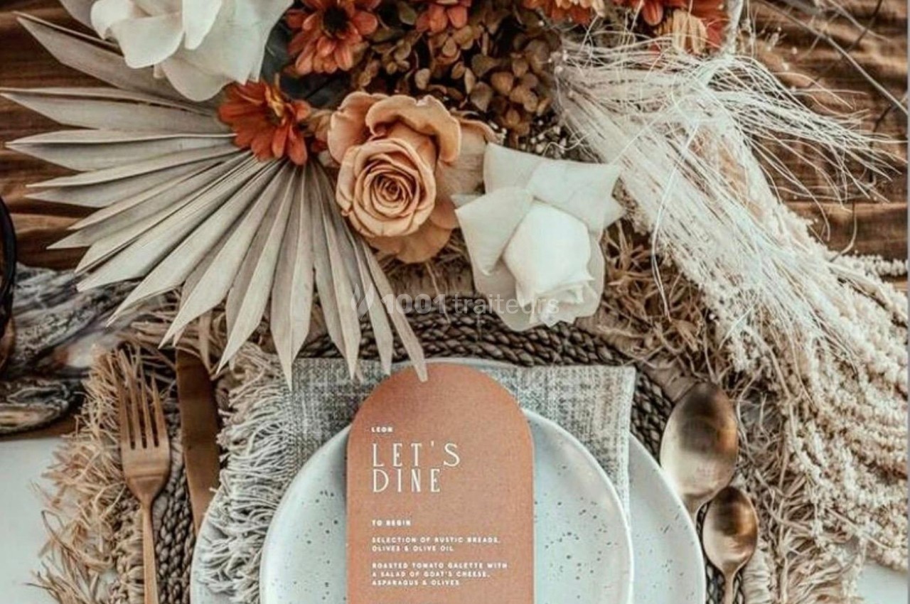 Table dressée avec des assiettes blanches, couverts dorés, menu beige et décoration florale aux tons neutres et orangés.