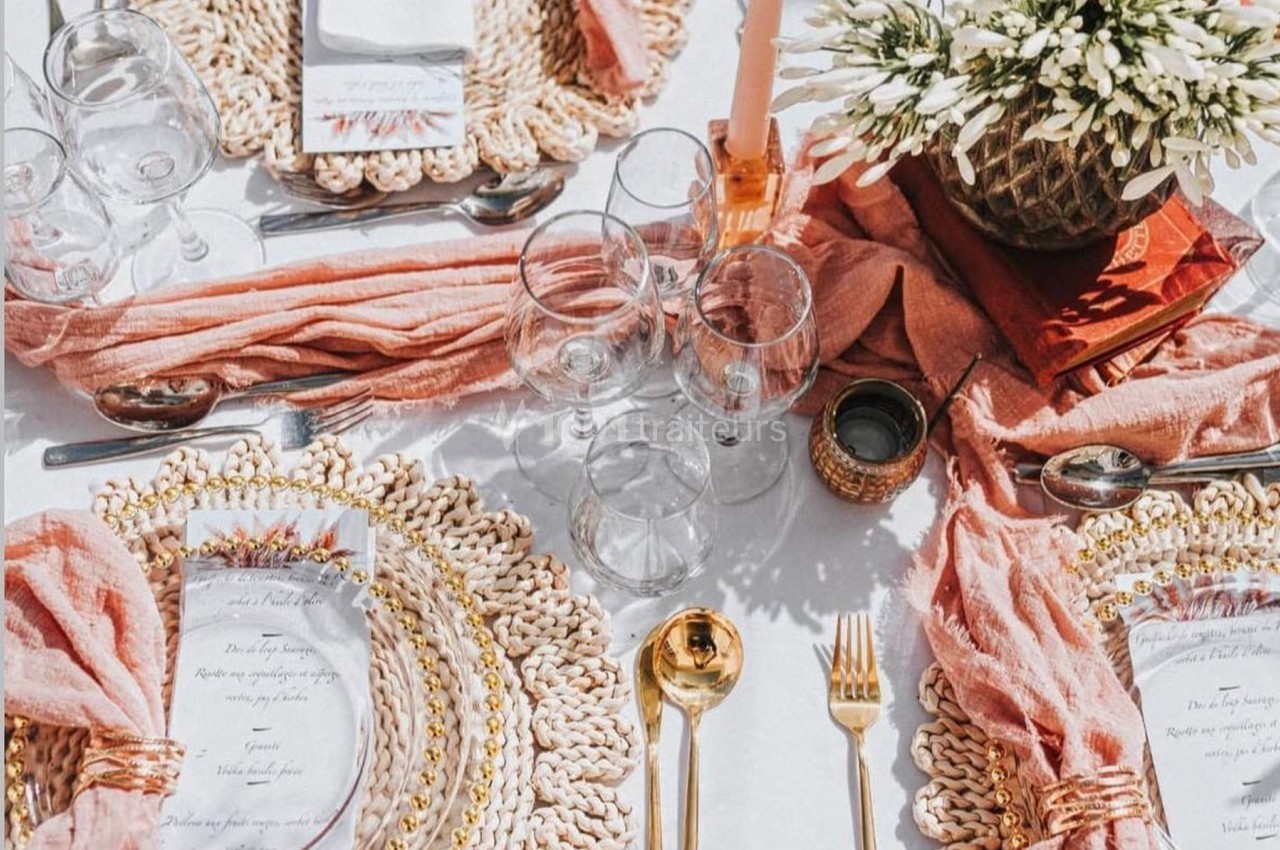 Table dressée avec des assiettes sur sets en rotin, couverts dorés, serviettes roses et centre de table floral.