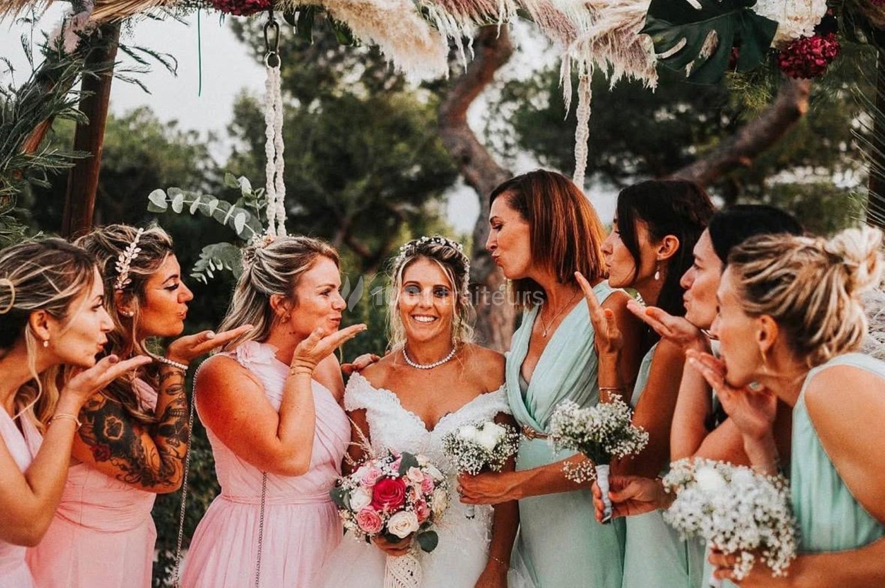 Des femmes en robes pastel entourent une mariée souriante sous une arche décorée de fleurs et de feuillages.
