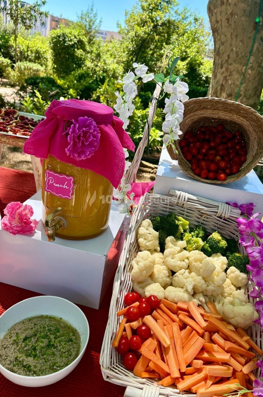 Distributeur de jus décoré, légumes crus en paniers, bol de sauce et panier de cerises sur une table en extérieur.