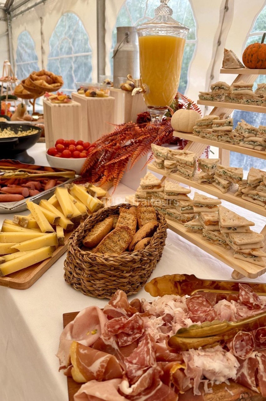 Buffet composé de charcuterie, fromages, pains, sandwichs, jus d'orange et décorations automnales sur une table blanche.