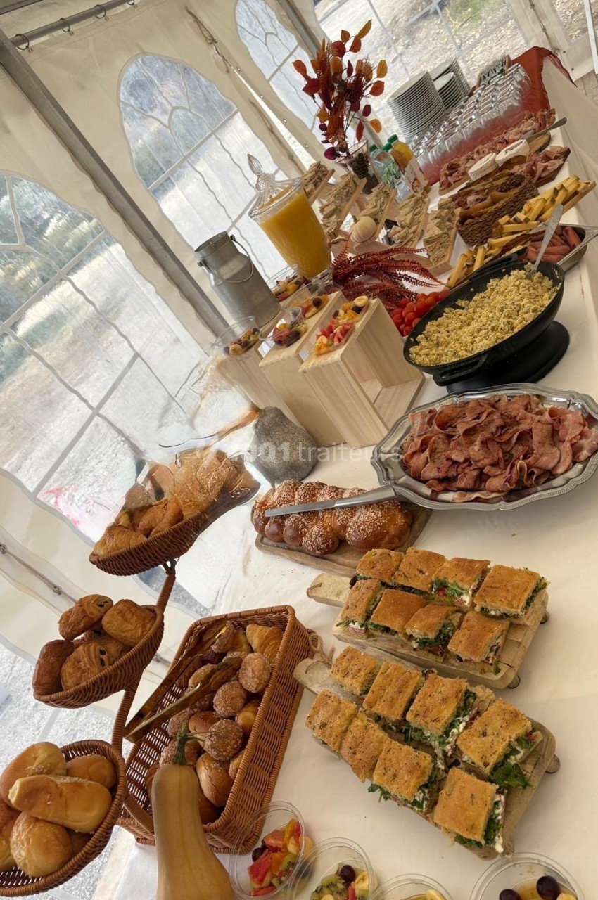 Buffet varié sous une tente, avec pains, viennoiseries, charcuterie, salades, desserts et boissons disposés sur des tables.
