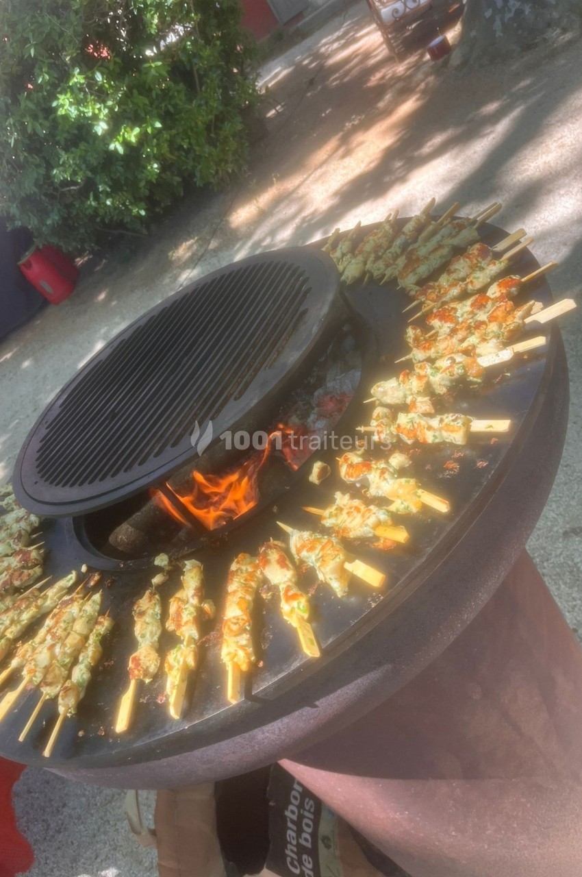 Brochettes de viande et légumes grillant sur un barbecue circulaire avec un feu de bois au centre.